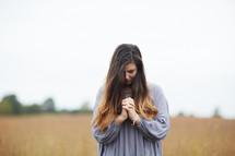 a woman standing in a field with head bowed and praying hands 