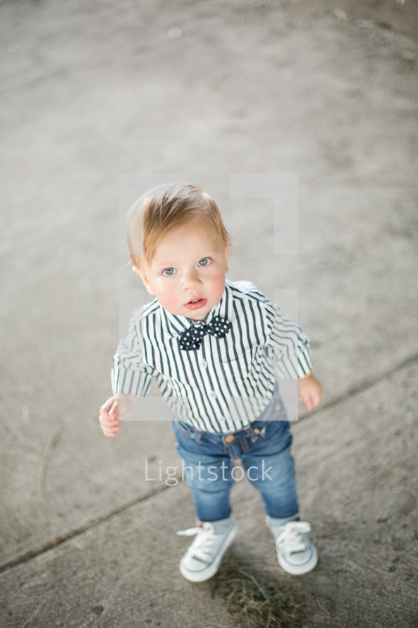 Toddler boy in a bow tie standing on the sidewalk outside.
