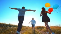 Happy family playing catch-up:child is running in front in center and his parents are running on sides of field holding colored balloons in hands. Outdoor games in summer leisure activity together.