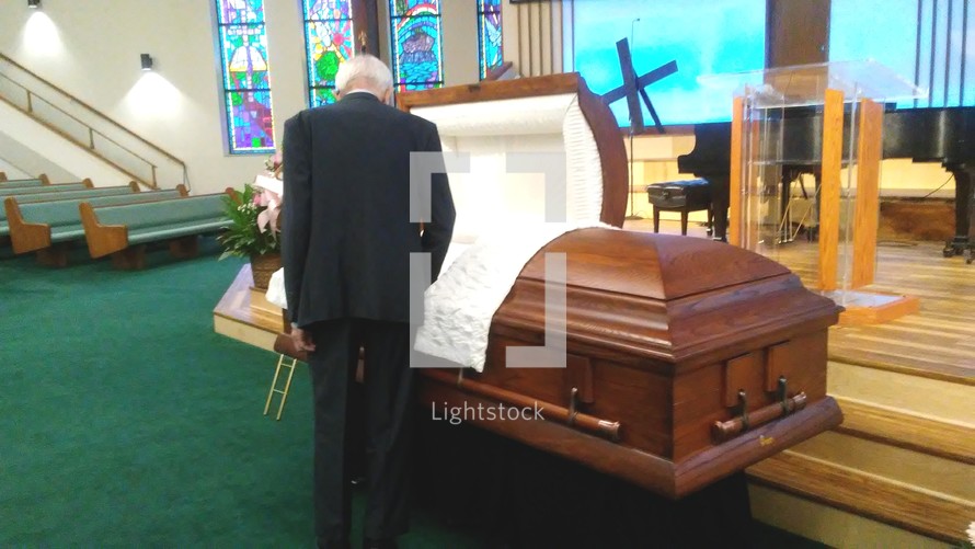 An elderly man with white hair and a black suit attends an open casket viewing at a funeral memorial service for a dearly departed loved one at a local church surrounded by open pews, stained glass windows and a cross appearing on the stage in the background. 