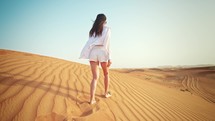 Rear view young woman walking on sandy mountain in desert with her arms outstretched. Wind blows her hair and jacket beautifully. Feeling freedom and happiness. Traveling in hot climate in Dubai.