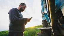 Man in field with phone in his hands stands by his tractor and knocks on wheel, checking condition of machine to diagnose serviceability of equipment. Running a small business selling grown products.