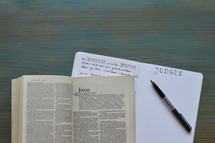 bible studies with a bible open at the book of Judges with notes and pen on a wooden cyan desk