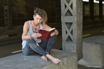 a punk woman sitting under a bridge reading a Bible 