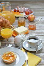 breakfast table with lots of fresh food like coffee, rolls, cheese, eggs, orange juice, tea, jam, butter, banana, apple, orange and a basket full of croissant and rolls