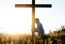 a man standing in a field next to a cross with head bowed 
