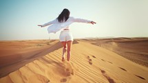 Rear view young woman walking on sandy mountain in desert with her arms outstretched. Wind blows her hair and jacket beautifully. Feeling freedom and happiness. Traveling in hot climate in Dubai.