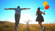 Happy family playing catch-up:child is running in front in center and his parents are running on sides of field holding colored balloons in hands. Outdoor games in summer leisure activity together.