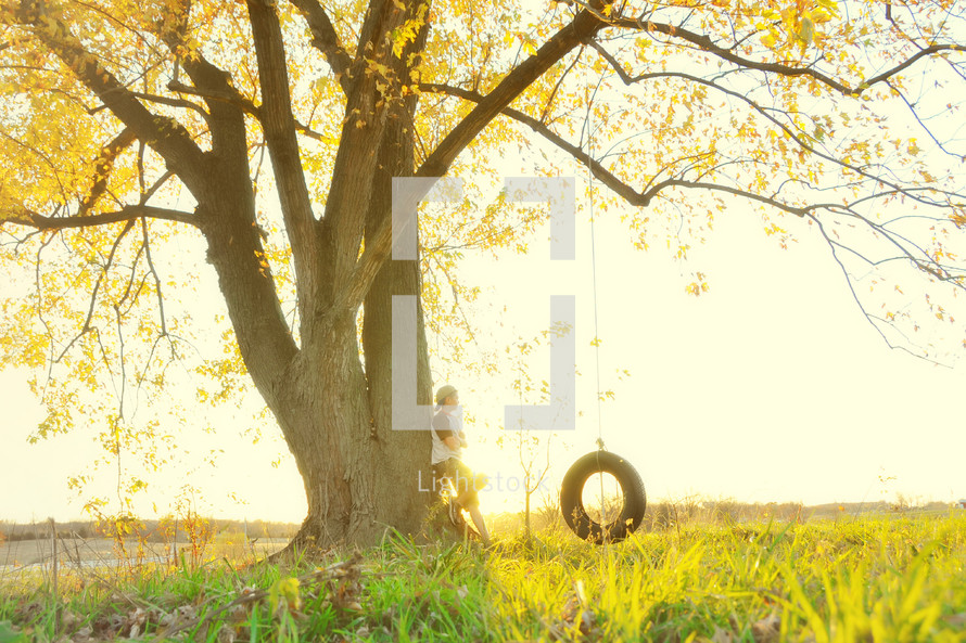 A person leaning against a tree near a tire swing.