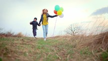 Happy sister and brother with with colorful balloons run across the field at sunset. Children play with balloons in the park. Happy kids on the nature with balloons. Freedom concept.