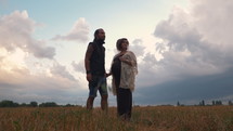 Man in a black cloth and a pregnant woman in a black long dress and dreadlocks embrace and are kept in hand, against the background of yellow grass field, nature, autumn or summer