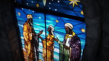 Close-up shot of a beautiful, dimly back-lit stained glass window of Nativity Wisemen with snow just starting to fall. Stained glass was generated with AI and composited into a 3D CGI scene.