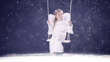 Young girl dressed as an angel, swinging, and smiling while it is snowing.