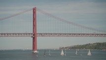 Regatta sailboats sailing beneath the iconic 25th of April Bridge in Lisbon, Portugal.