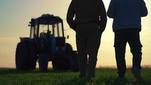 Teamwork concept. Silhouette two male farmers walking in a green field against sunset. Team farmers stand in a field on the background of agricultural machinery. Agronomists discuss harvest.