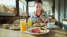 Young woman eats breakfast in the wooden cafe on mountain, alpine view, snow on hills. Breakfast on terrace of cafe in mountain resort. Healthy lifestyle, travel, family moments together concept.