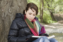 woman reading a Bible resting against a tree outdoors