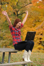 A woman on a picnic table with a laptop computer on her lap and arms raised.