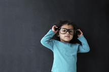 toddler girl wearing reading glasses against a chalkboard background.