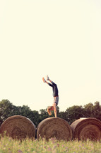 Woman doing a handstand on a rolled bale of hay in a field.