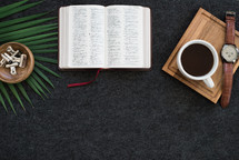 open Bible, tray, coffee mug, palm fronds, and watch on a desk 