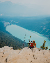 Woman sitting on a rock cliff looking out at a scenic mountain lake view.
