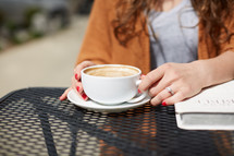 a woman sitting at an outdoor table with a Bible, drinking coffee 