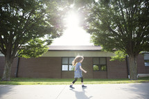 A little girl walking down the sidewalk next to school.