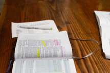 Open Bible on wooden table, next to paper and highlighter, yellow and pink highlighter on Bible verses