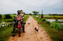 family on a dirt bike on a dirt road and chickens 