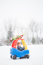 a toddler boy in a toy car with a Christmas tree on top 