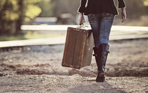 a woman walking on a dirt road carrying a suitcase 