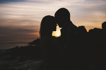 man and woman kissing on a beach in front of the ocean at sunset