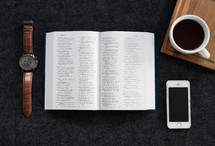 An open Bible, wristwatch, cell phone and a cup of coffee on a black surface.