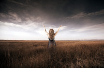 woman standing in a field  under a stormy sky with her arms raised in worship 