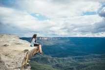 Woman sitting on the edge of a cliff overlooking a valley.