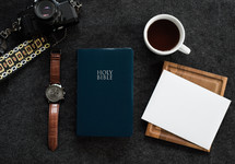A Bible, wristwatch, cup of coffee, camera and a blank envelope on a black background.