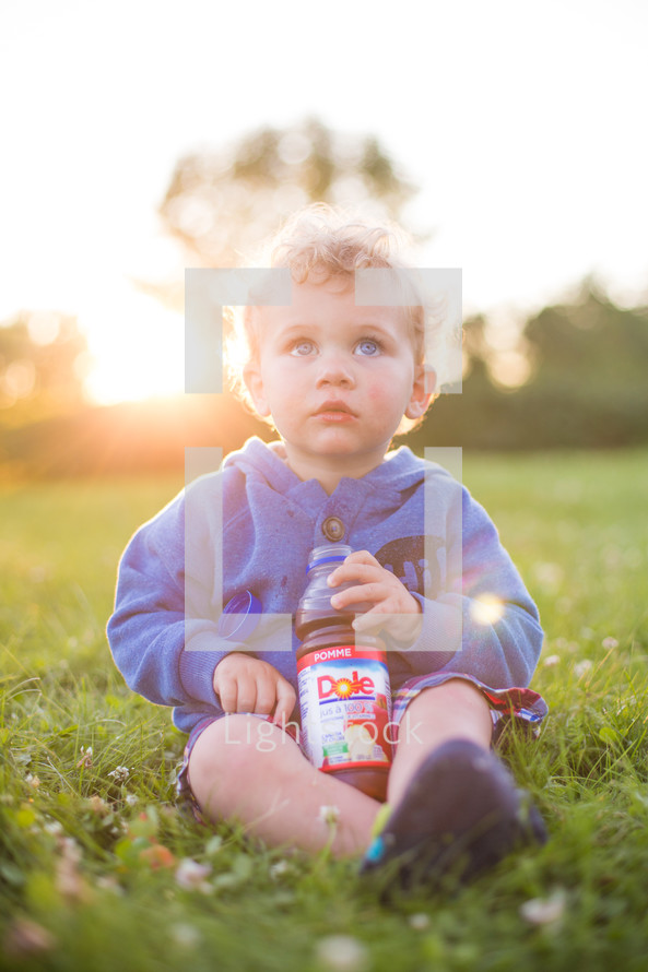 A little boy sitting in green grass with a bottle of juice.