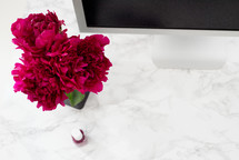 A computer monitor, dark pink flowers and a bottle of nail polish on a white granite surface.