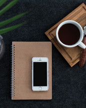 A cell phone on a spiral notebook next to a cup of coffee and wristwatch on a wooden board.