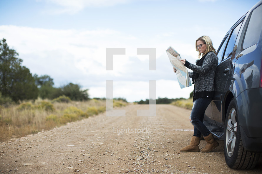 A woman standing by her car on an empty road looking at a road map.