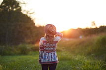 a woman in a sweater and a hat standing in the middle of a field at sunset 