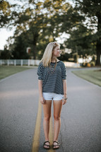 A young blonde woman in shorts standing on the yellow stripe in the middle of a road.