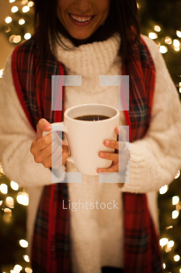 a woman holding a mug of hot cocoa standing in front of a Christmas tree 