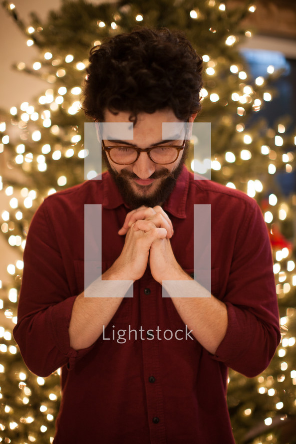 a man with head bowed in prayer standing in front of Christmas tree 