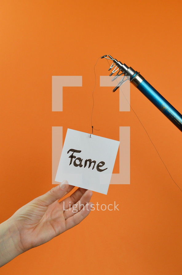 a hand grabbing for a piece of paper with the word FAME on it hanging from a fishing line