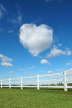 Heart-shaped cloud over fence rails in a grassy pasture.