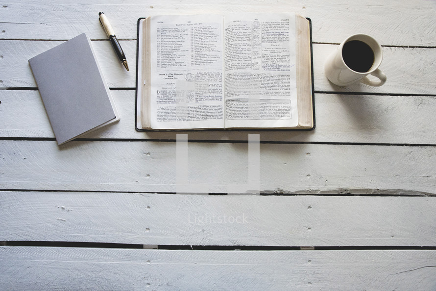 pages of a Bible, pen, journal, and coffee cup on a white wood background 