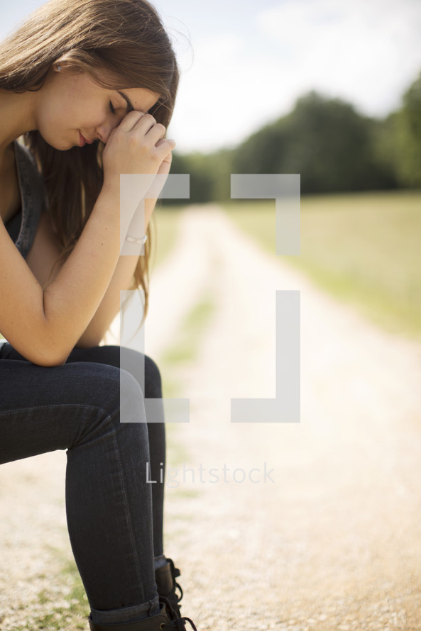 woman sitting on a suitcase on a dirt road with head bowed in prayer 