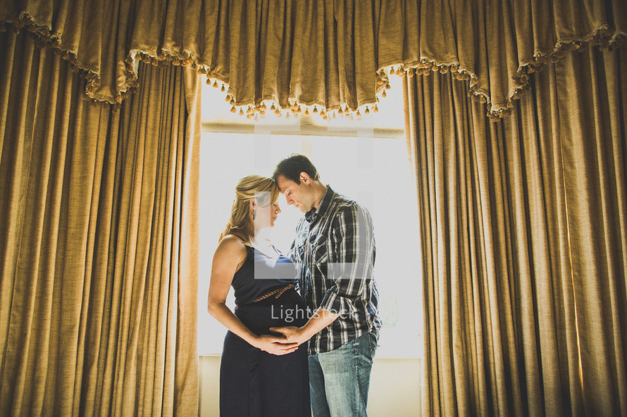 Pregnant couple embracing in front of window with thick drapes.